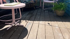 Eine Holzterrasse mit einem Korussofa, zwei rosa Stühlen, einem kleinen Tisch und einem Blumentopf.