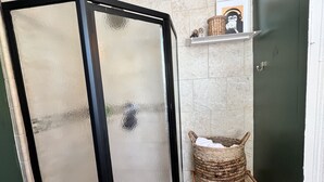 Eine Duschkabine mit Milchglas-Türen, ein Korbwäscher auf einem Hocker und eine Ablage mit Dekorationselementen.