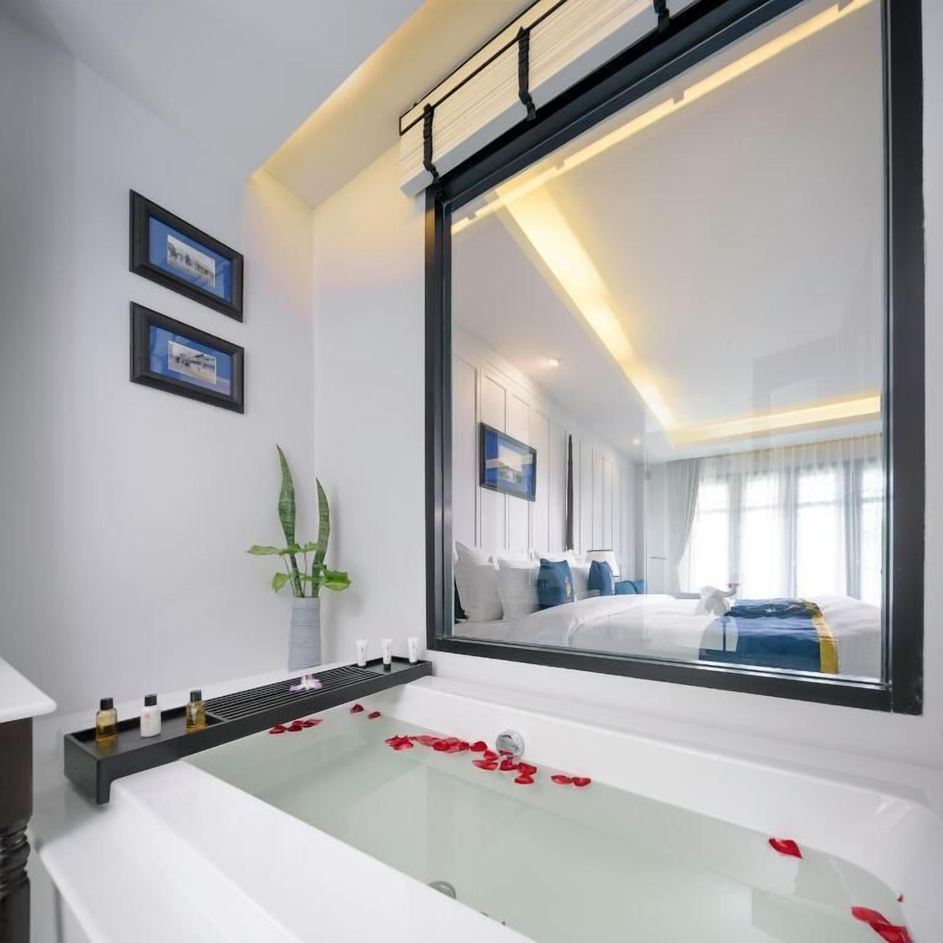 A modern hotel room with a large bathtub filled with rose petals, a bedside table with toiletries, a large mirror reflecting the room, and a potted plant on the windowsill.