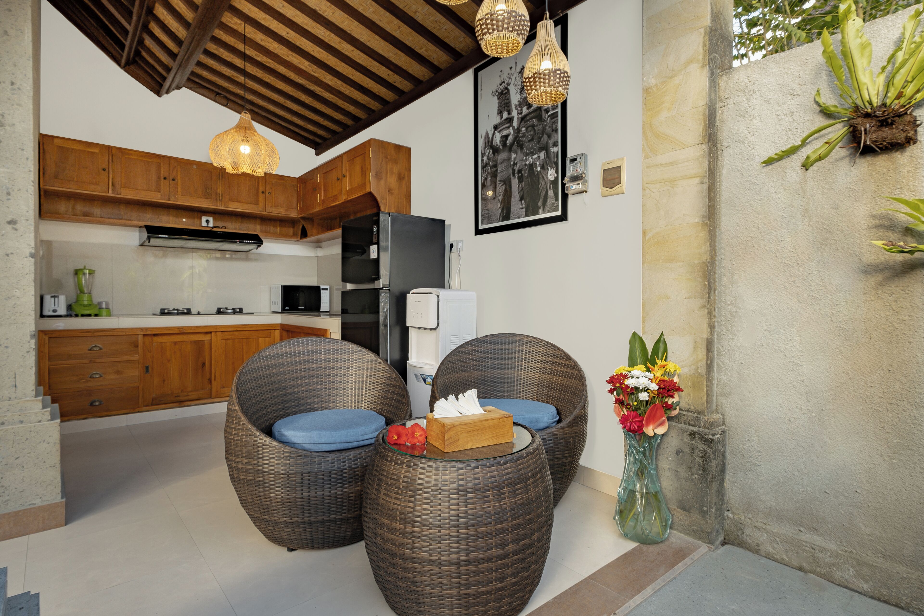 A kitchen with wooden cabinets, a refrigerator, and a microwave. There are wicker chairs with cushions and a vase with flowers.