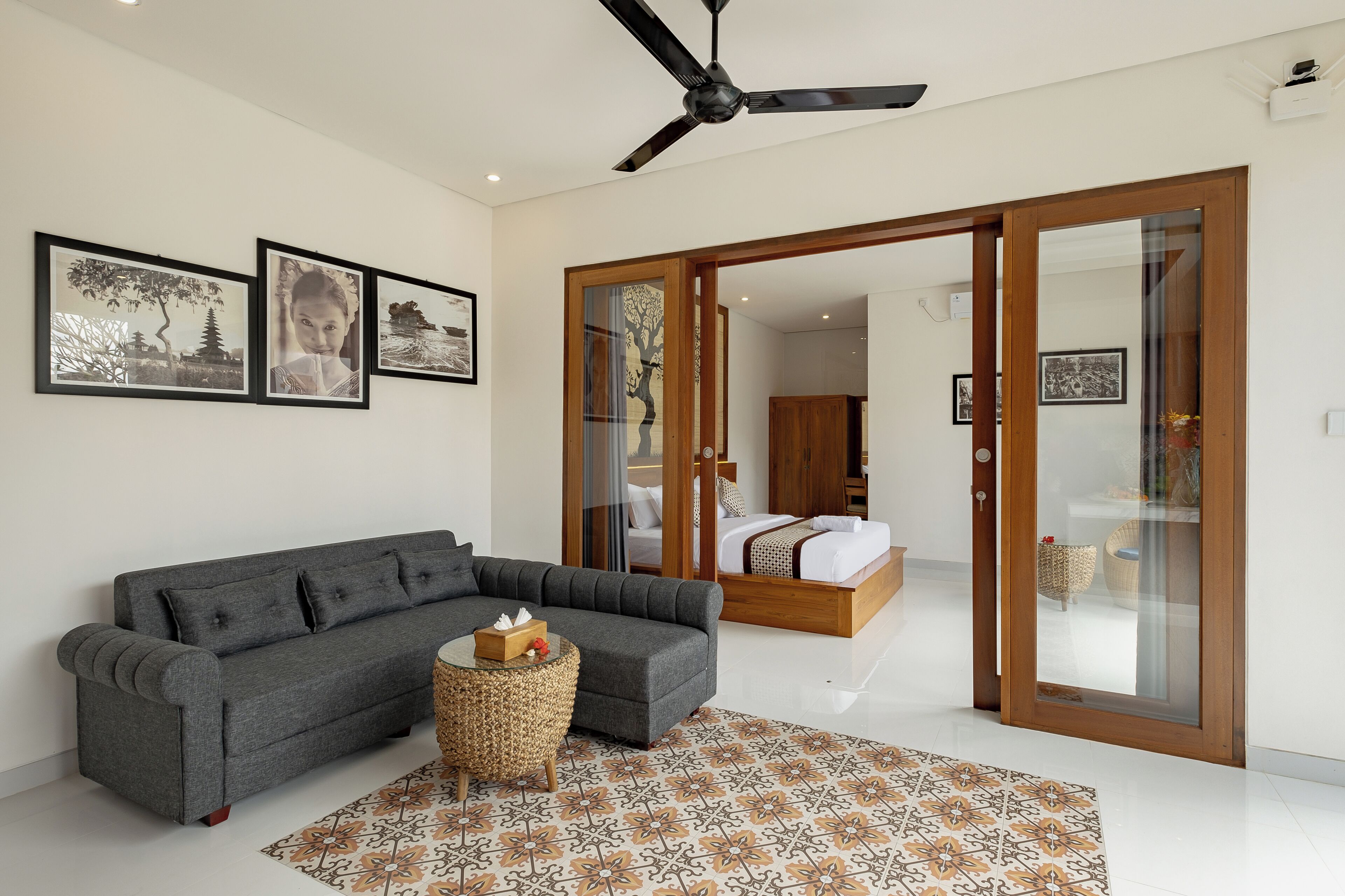 A modern living room with a grey sofa, a small round table, and a patterned rug. There is a ceiling fan and a bed with a wooden frame visible through an open door.