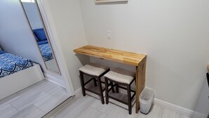 Eine kleine Küchenzeile mit Holzausstattung, zwei Barhocker, einem Mülleimer und einem Bett mit blauer Bettwäsche.