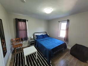 Ein Schlafzimmer mit Bett, Nachttisch, Sessel, kleinem Tisch, Fenster mit Vorhängen und Deckenleuchte.