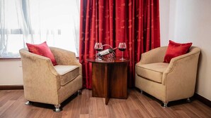 Une pièce avec deux fauteuils beiges, une petite table en bois sur laquelle se trouvent des verres à vin et une bouteille, des rideaux rouges et une fenêtre ornée de rideaux légers.