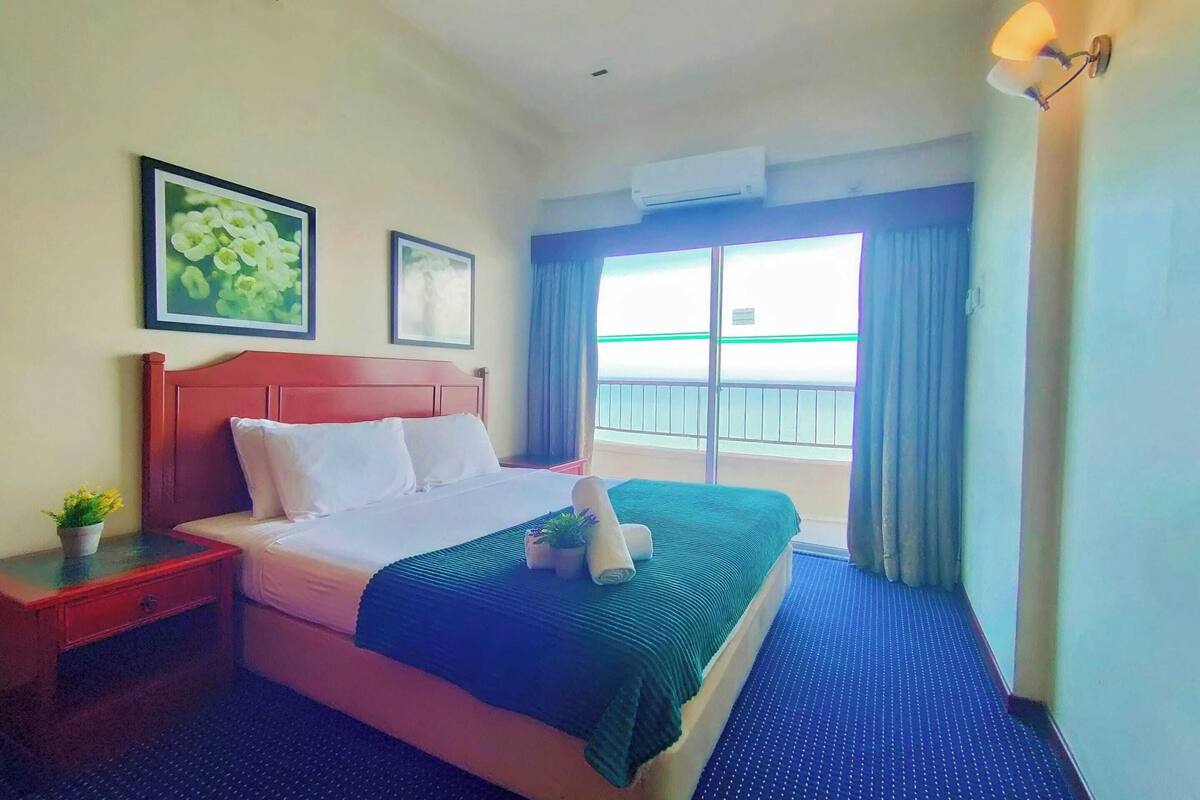 A hotel room with a bed, a nightstand, a potted plant, a window with curtains, and a view of the outside.