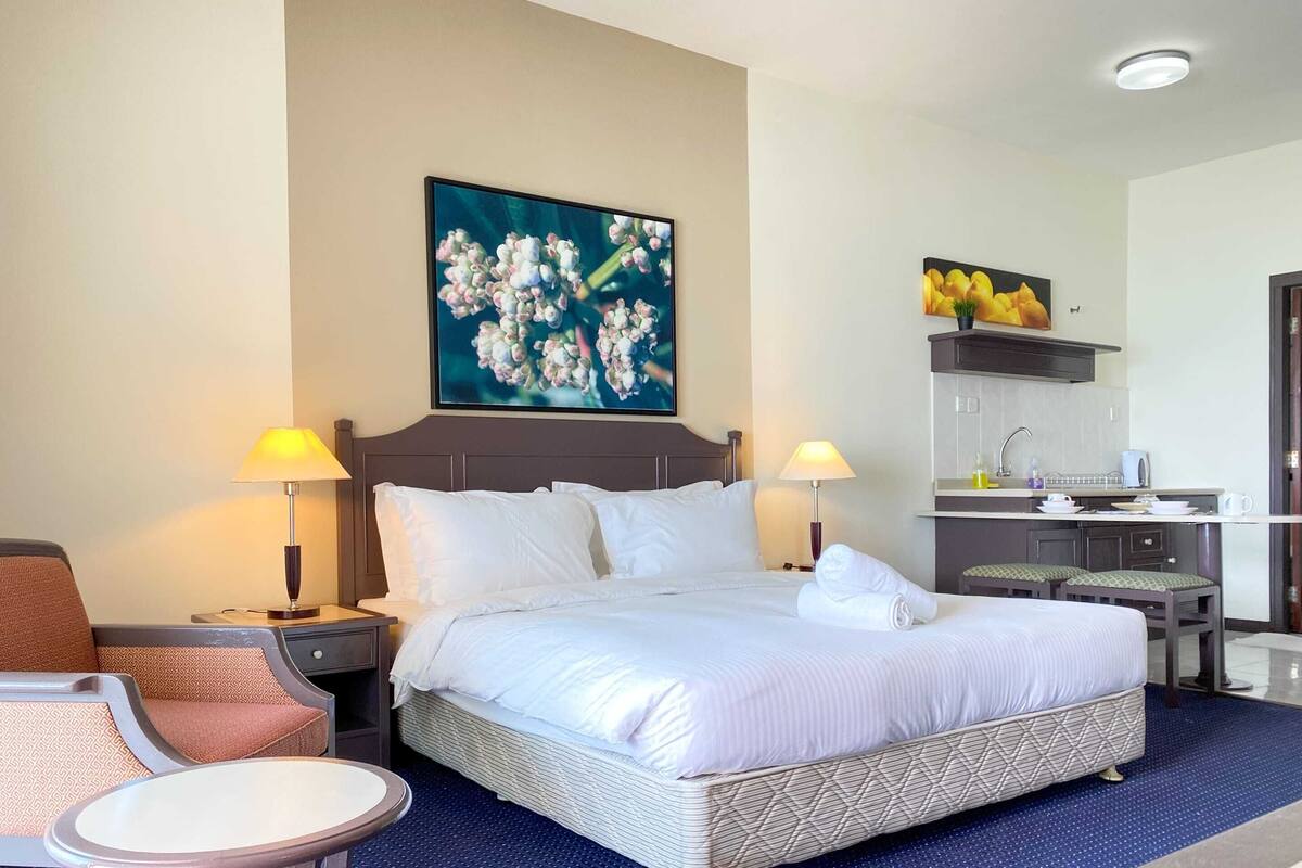A hotel room with a large bed, two bedside lamps, a desk with a chair, a small kitchenette, and a painting on the wall.