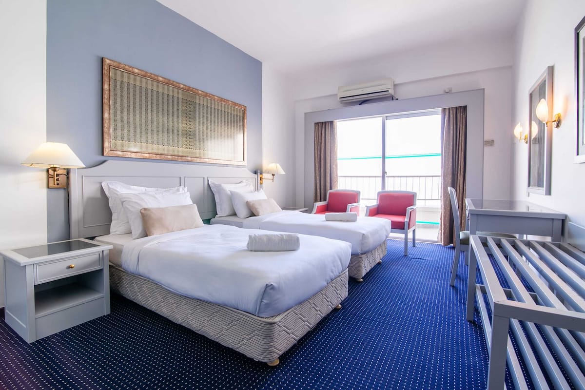 A hotel room with two beds, a small desk, a chair, a large window, and a balcony.