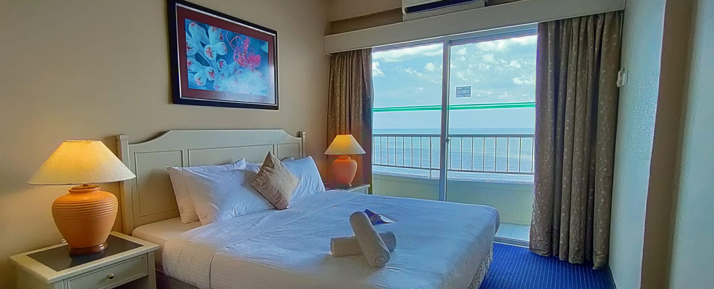 Habitación de hotel con cama, dos lámparas de noche, un cuadro en la pared y vista al mar a través de un ventanal amplio.