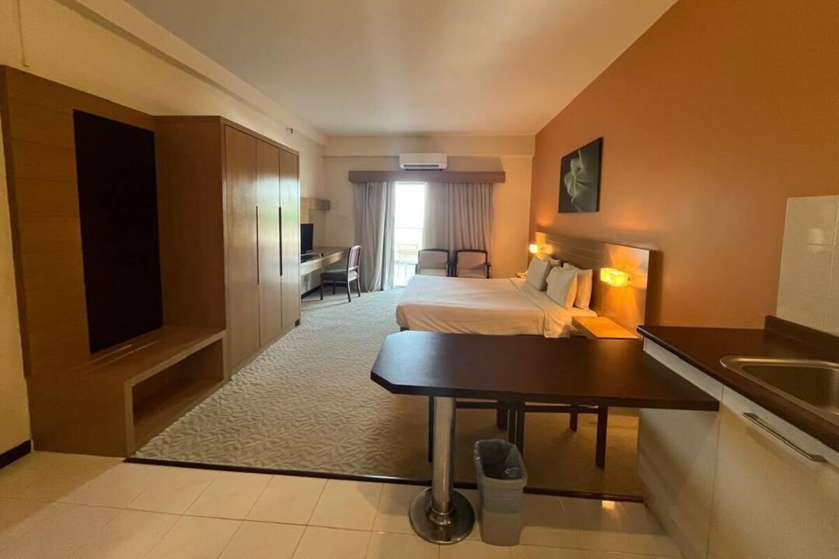A hotel room with a bed, bedside table, lamp, and a small kitchenette.