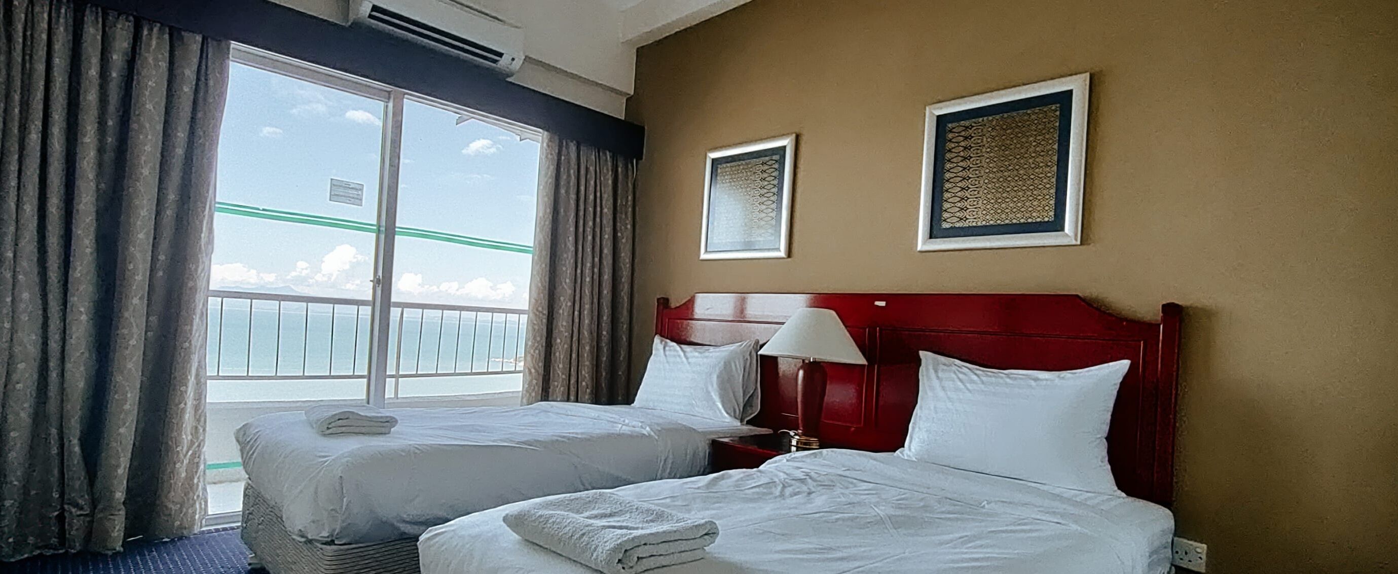 Habitación de hotel con dos camas, un ventanal grande con cortinas, cabecera roja y dos cuadros en la pared.