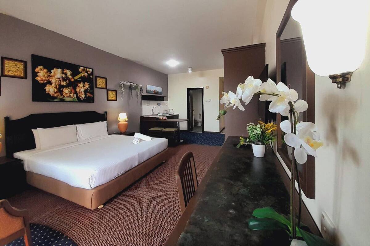 A hotel room with a bed, a desk, a chair, a small kitchenette, and a bathroom visible in the background.