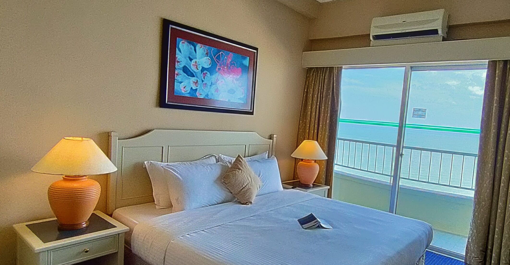 A hotel room with a bed, two bedside lamps, a framed abstract painting, and a view of a balcony with a railing.