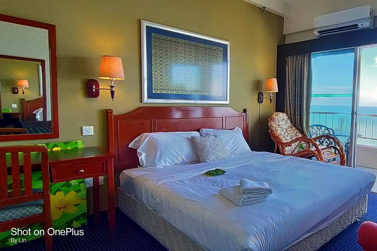 A hotel room with a bed, a chair, a desk, a mirror, a window with a view of the sea, and a vase with flowers.