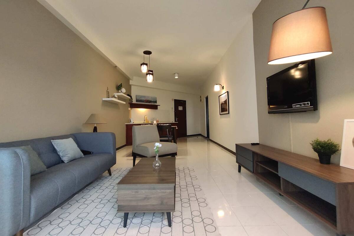 A modern living room with a sofa, coffee table, and TV. There is a kitchen in the background.