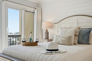 A bed with a white quilted bedspread, a white hat, a wicker basket with glasses and a bottle, a nightstand with a lamp, and a view through a window. - Oceanfront, By 30A, Pool, Sauna, Boat Slips FREE! (Panama City Beach)