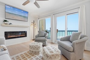 A living room with a fireplace, two grey armchairs, a coffee table, and a large window with a view of the ocean. - Oceanfront, By 30A, Pool, Sauna, Boat Slips FREE! (Panama City Beach)