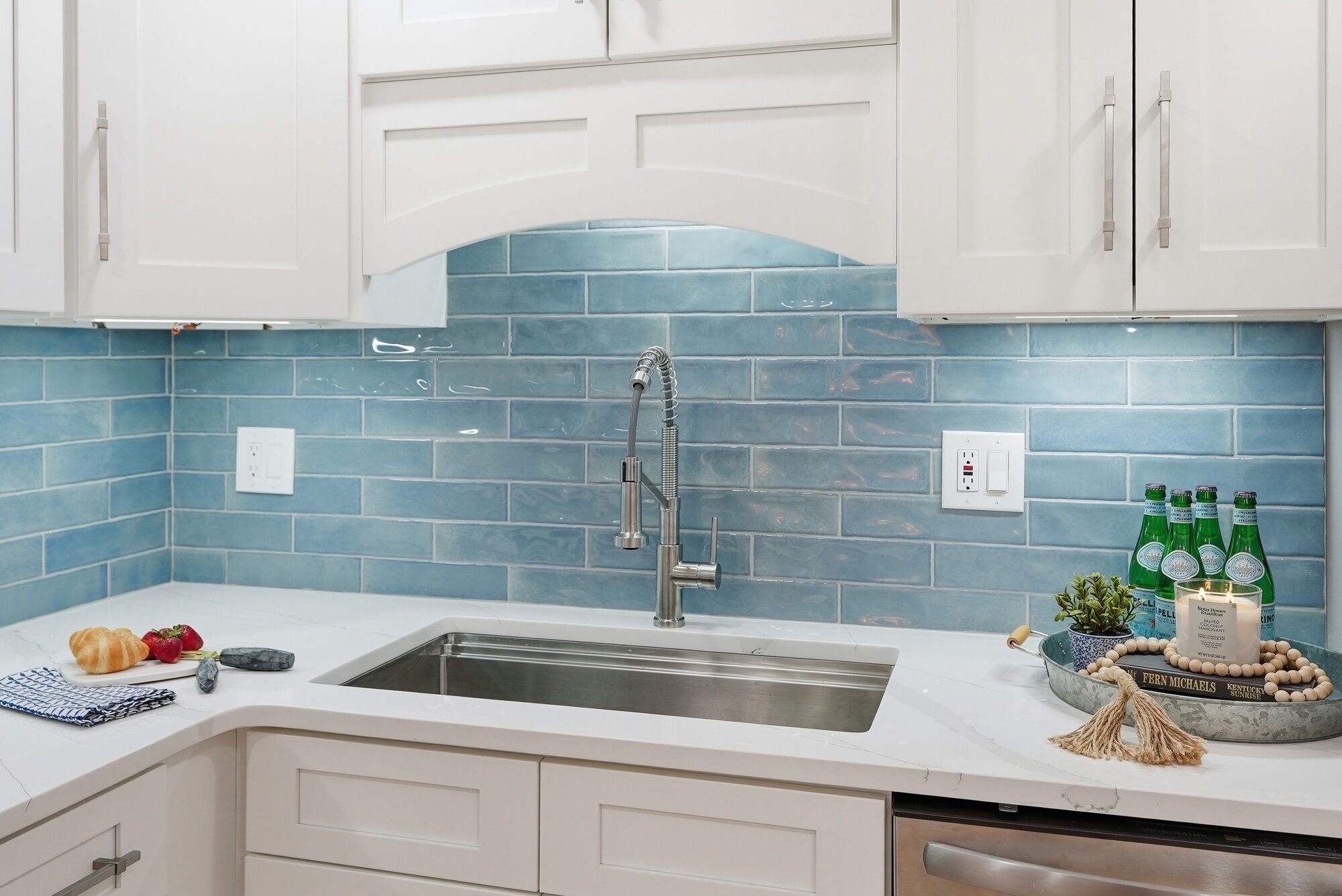 A kitchen with white cabinets, a blue-tiled backsplash, a stainless steel sink, and a countertop with various items.