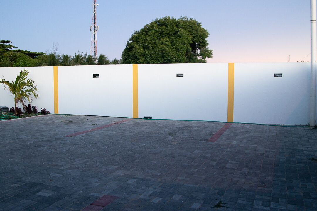 A paved outdoor area with a white wall featuring yellow vertical stripes and small plaques.