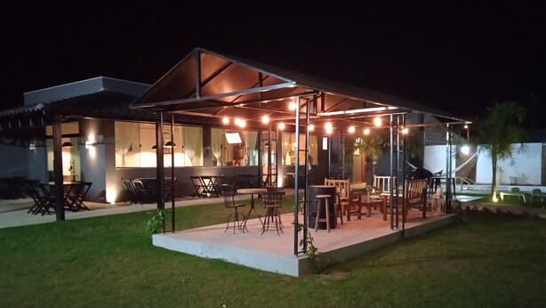 An outdoor dining area with tables and chairs, a covered seating area, and a building with large windows in the background. - Pousada Porto Prime (Sao Joao Da Barra)