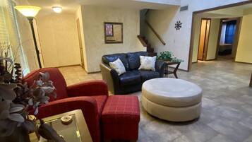 A spacious living area with a dark leather sofa, red armchair, and ottoman. There is a small table with a plant and a framed picture on the wall.