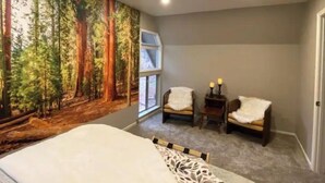 Ein Schlafzimmer mit Waldgemälde, einem Bett, zwei Sessel mit weißen Überwürfen, einem Nachttisch mit Kerzen und einem Fenster.