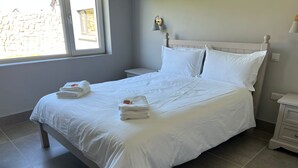 Ein ordentlich bezogenes Bett mit weißen Leinen und Handtüchern, ein Nachttisch, eine Wandlampe und ein Fenster mit Blick auf eine Steinmauer.