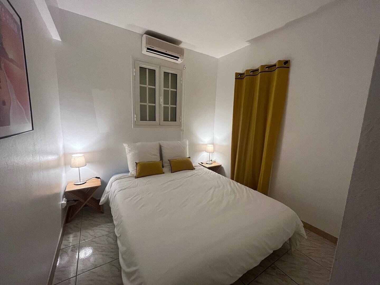 Ein Hotelzimmer mit einem Bett, zwei Nachttischlampen, einem Fenster mit Vorhängen und einer Klimaanlage.