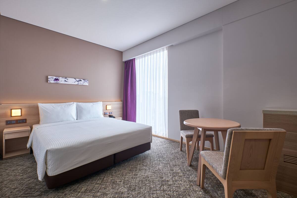A hotel room with a large bed, a bedside table with a lamp, a chair, a small round table, and a window with curtains.
