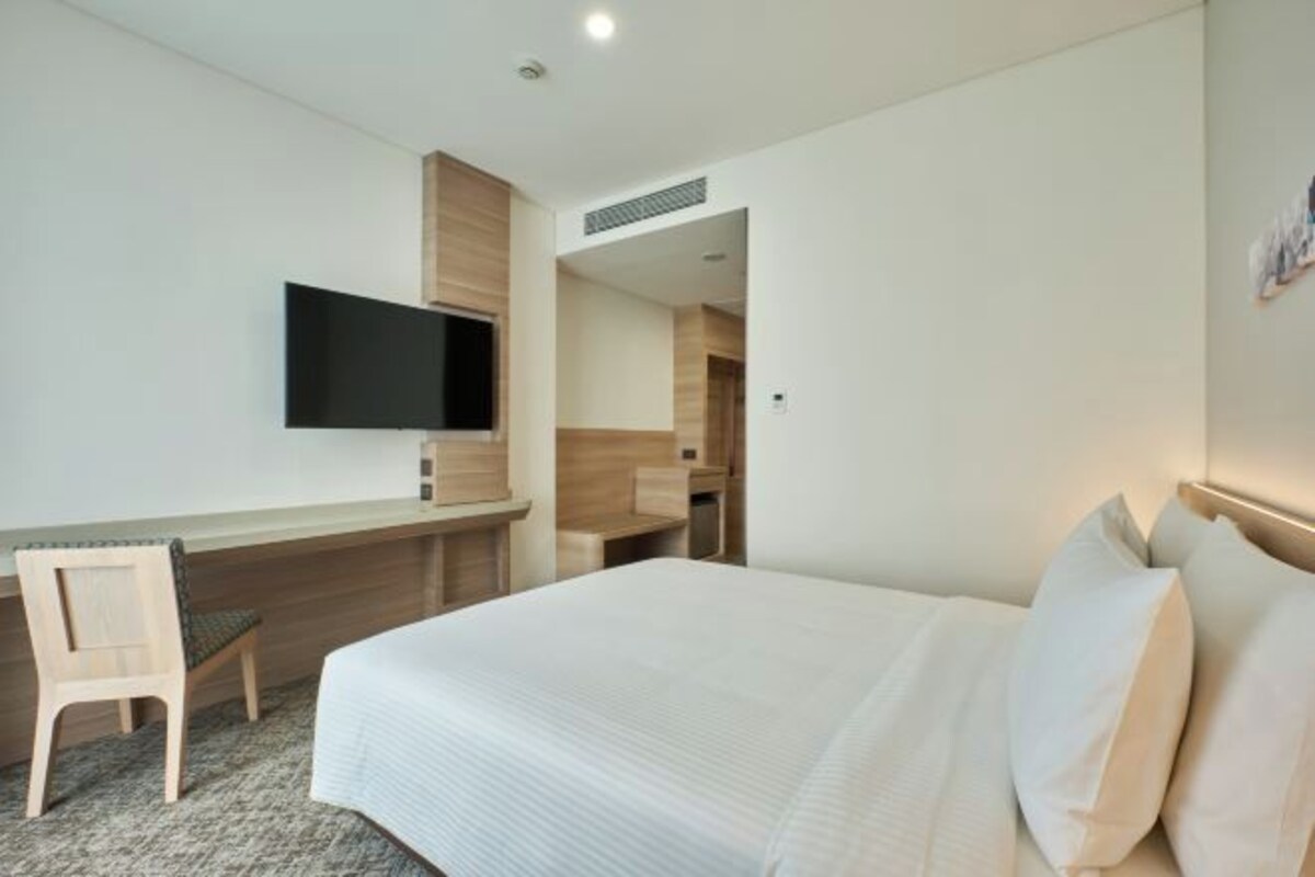 A hotel room with a bed, a desk, a chair, and a flat-screen TV mounted on the wall.