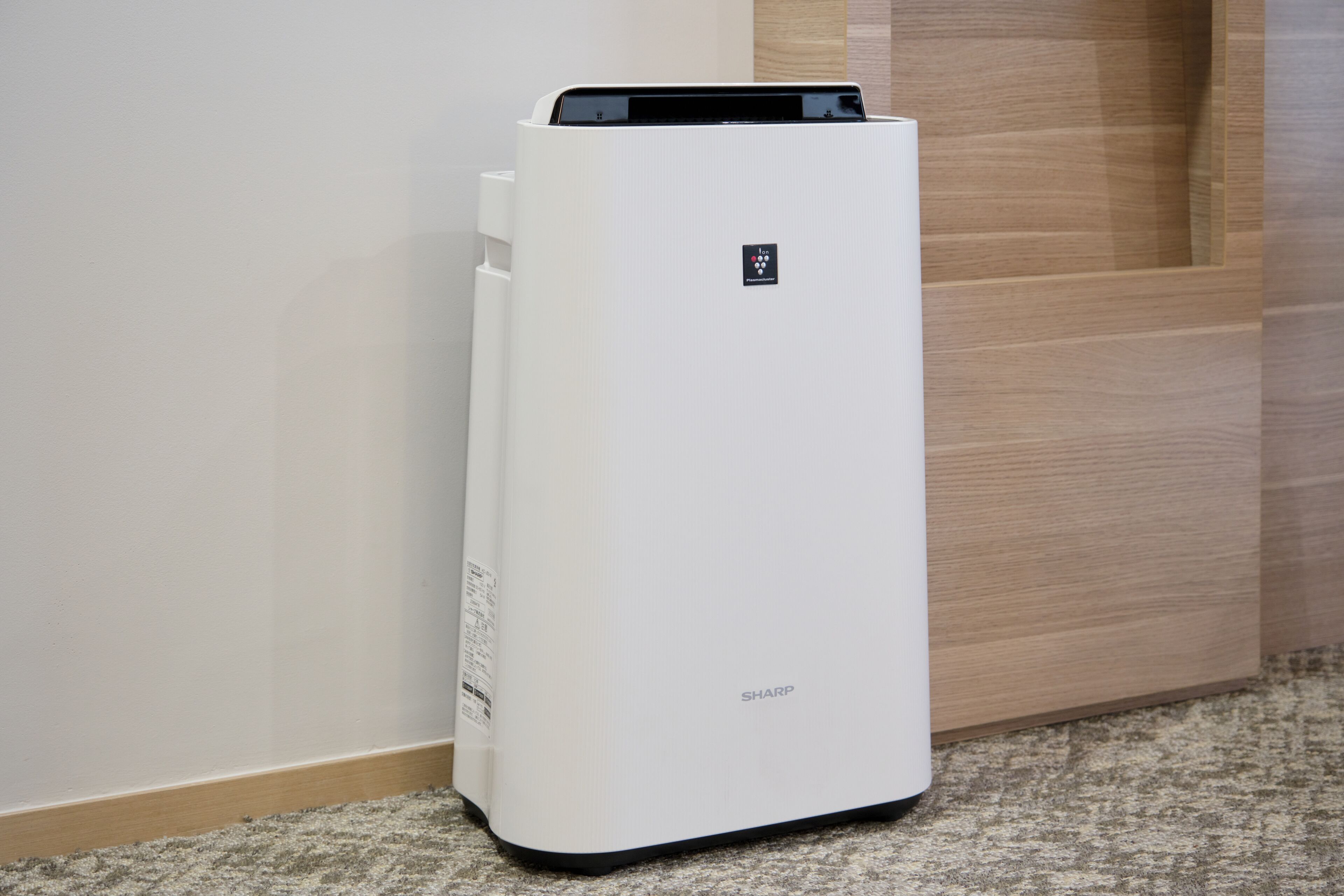 A white, modern air purifier with a control panel and a brand logo, placed on a carpeted floor next to a wooden door.