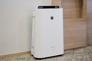 A white, modern air purifier with a control panel and a brand logo, placed on a carpeted floor next to a wooden door.