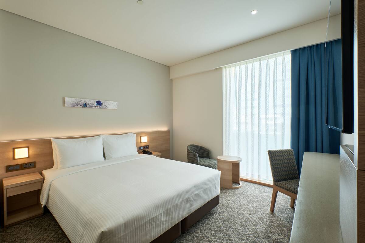 A hotel room with a large bed, two bedside tables, a chair, a small round table, and a large window with curtains.
