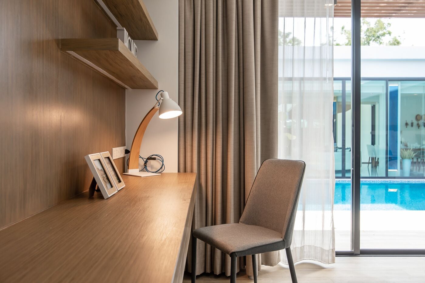 A modern room with a wooden desk, a chair, a desk lamp, and a view of a swimming pool through large windows.