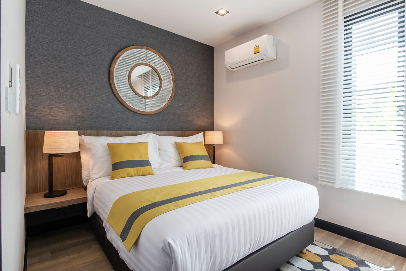 A modern hotel room with a bed, bedside lamps, a wall-mounted air conditioner, and a window with blinds.