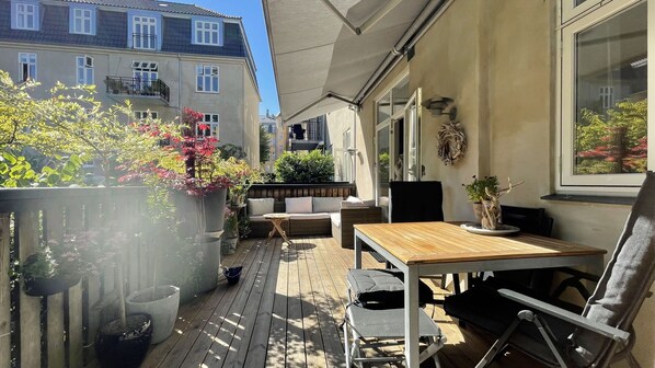 A wooden deck with outdoor furniture, potted plants, and a view of neighboring buildings.