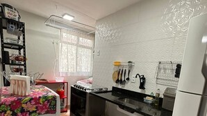 Eine kleine Küche mit Gasherd, Kühlschrank und Spüle. Ein Fenster mit Vorhang, ein Regal mit Küchenutensilien und ein kleiner Mülleimer.