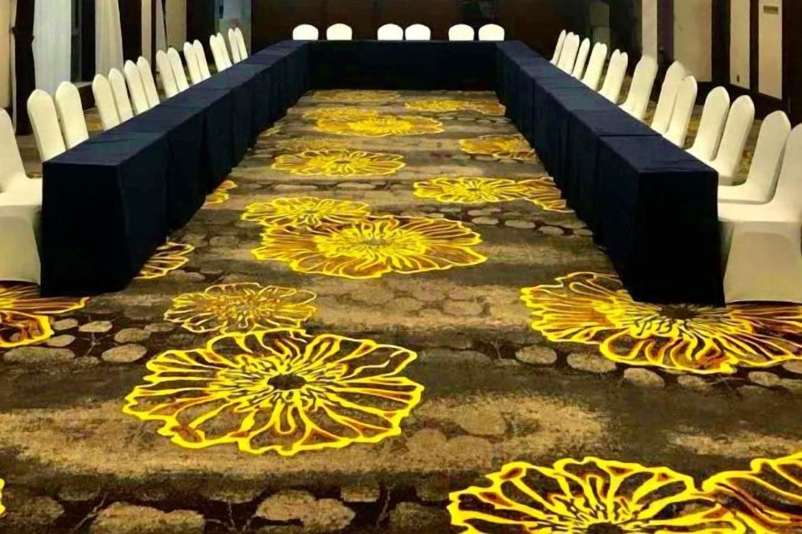 Sala de conferência com uma longa mesa, cadeiras brancas e um tapete com motivos florais.