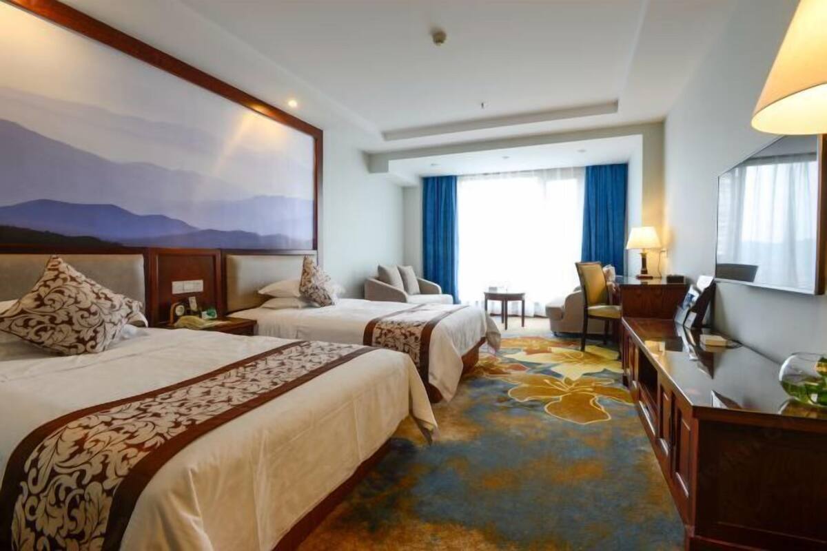 A hotel room with two beds, a desk, a chair, a large window with blue curtains, and a painting of mountains.