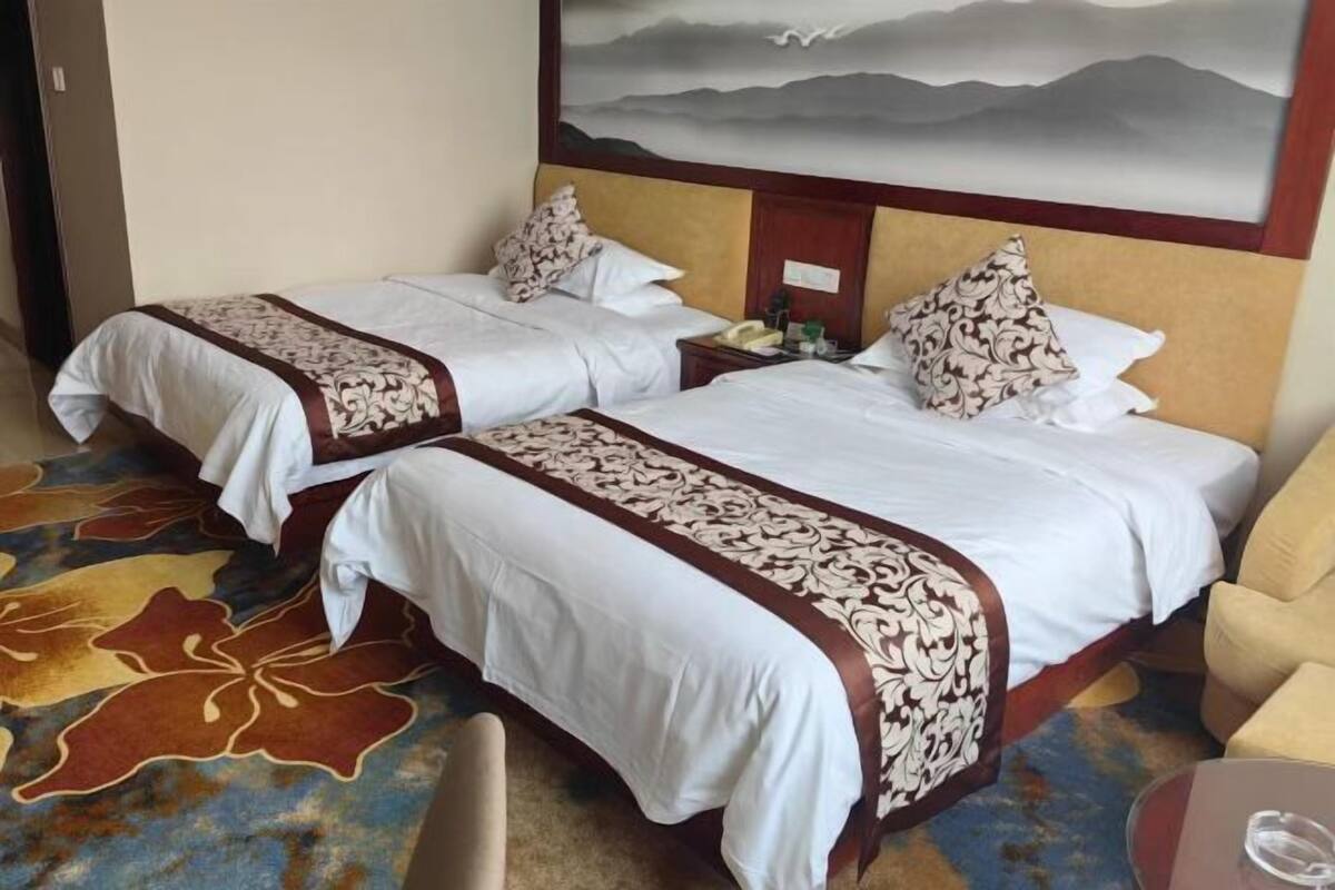 A hotel room with two beds, a large painting on the wall, a carpet with a floral pattern, and a chair.