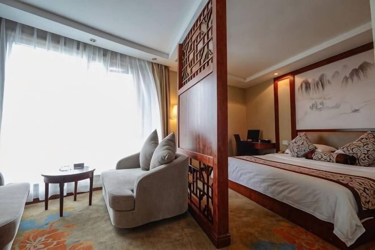 A hotel room with a bed, a chair, a small table, and a large window with curtains.