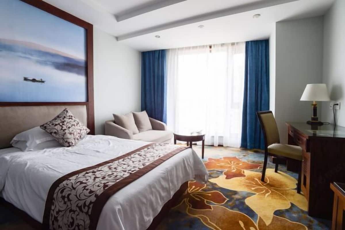 A hotel room with a bed, a desk, a chair, a small table, and a painting on the wall.