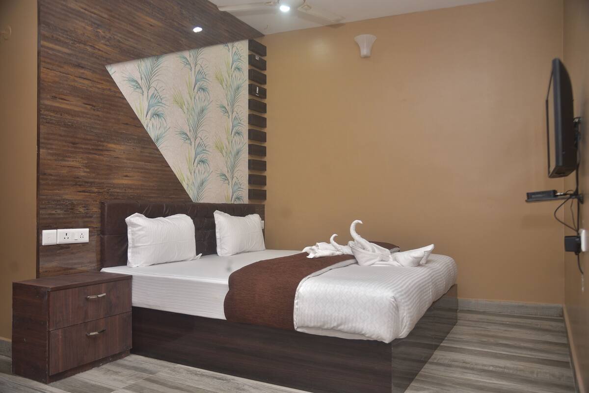 A modern hotel room with a wooden headboard, a large bed with white linens, a brown throw blanket, and a wall-mounted flat-screen TV.
