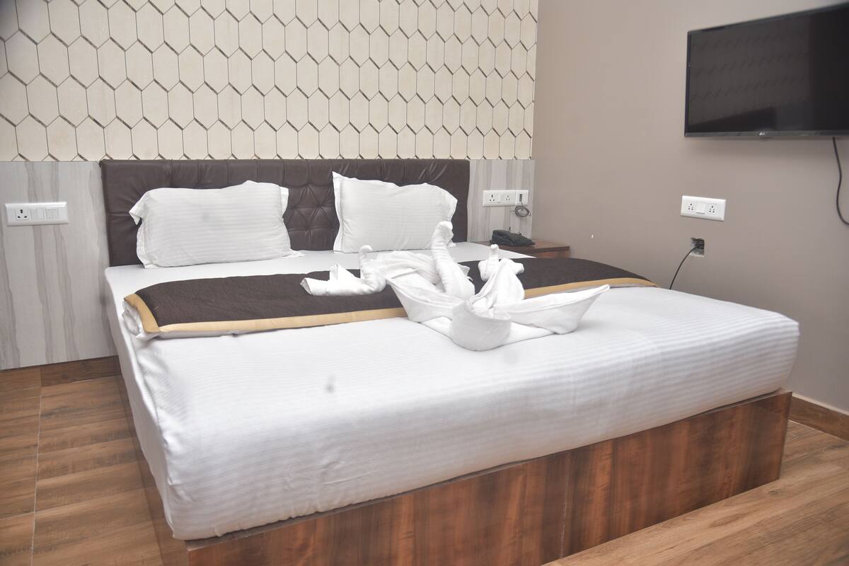 A neatly made bed with white linens and a brown headboard, a flat-screen TV mounted on the wall, and a wooden floor.