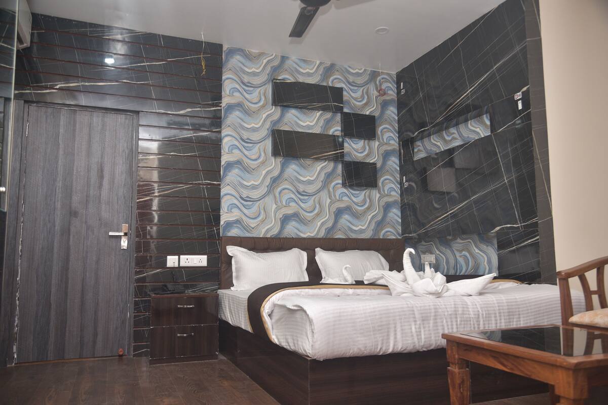 A modern hotel room with a large bed, a wooden nightstand, a dark wooden door, and a marble-patterned wall.