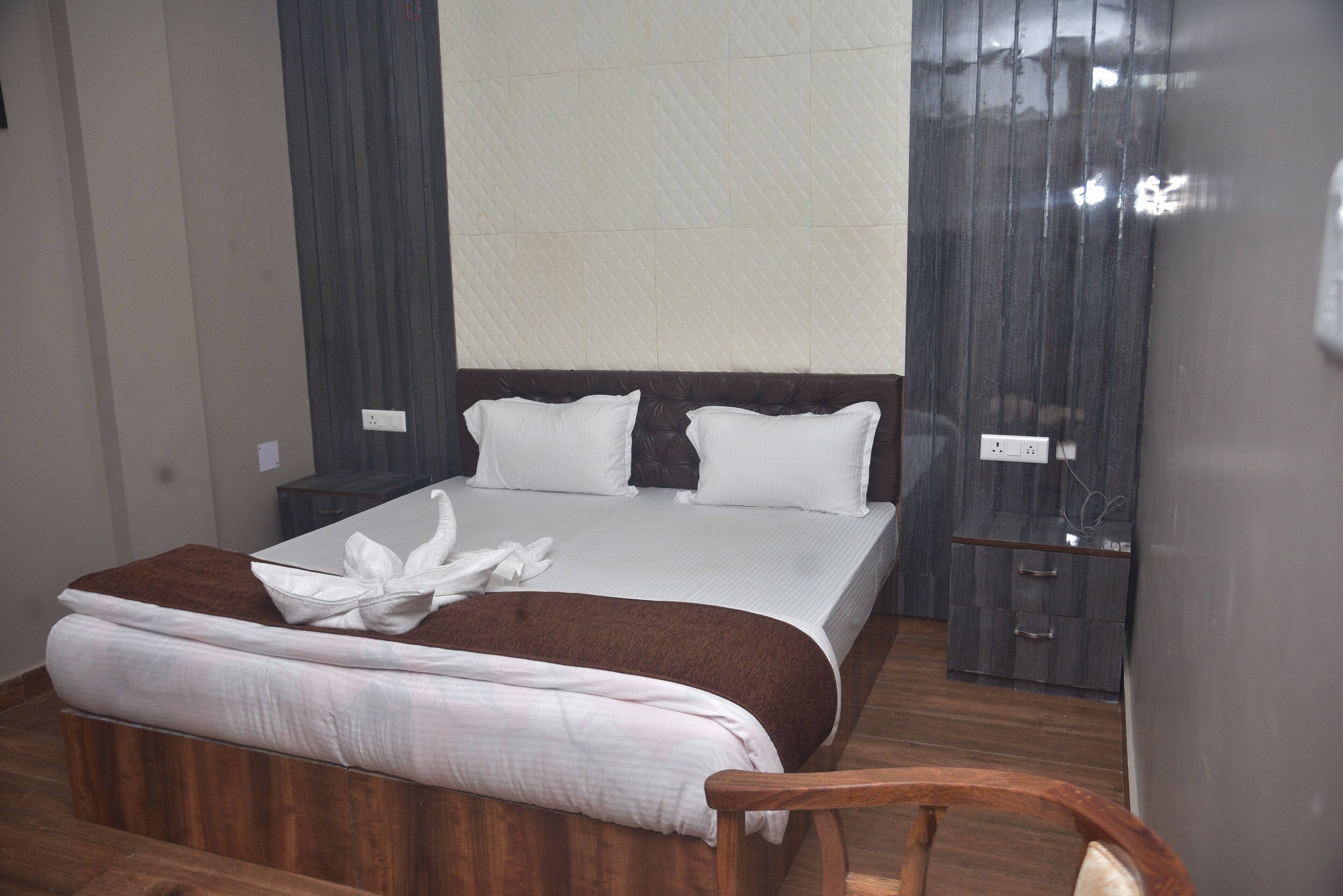 Een opgemaakt bed met witte lakens en een bruine plaid, een houten nachtkastje met een lamp, en een donker houten garderobe.
