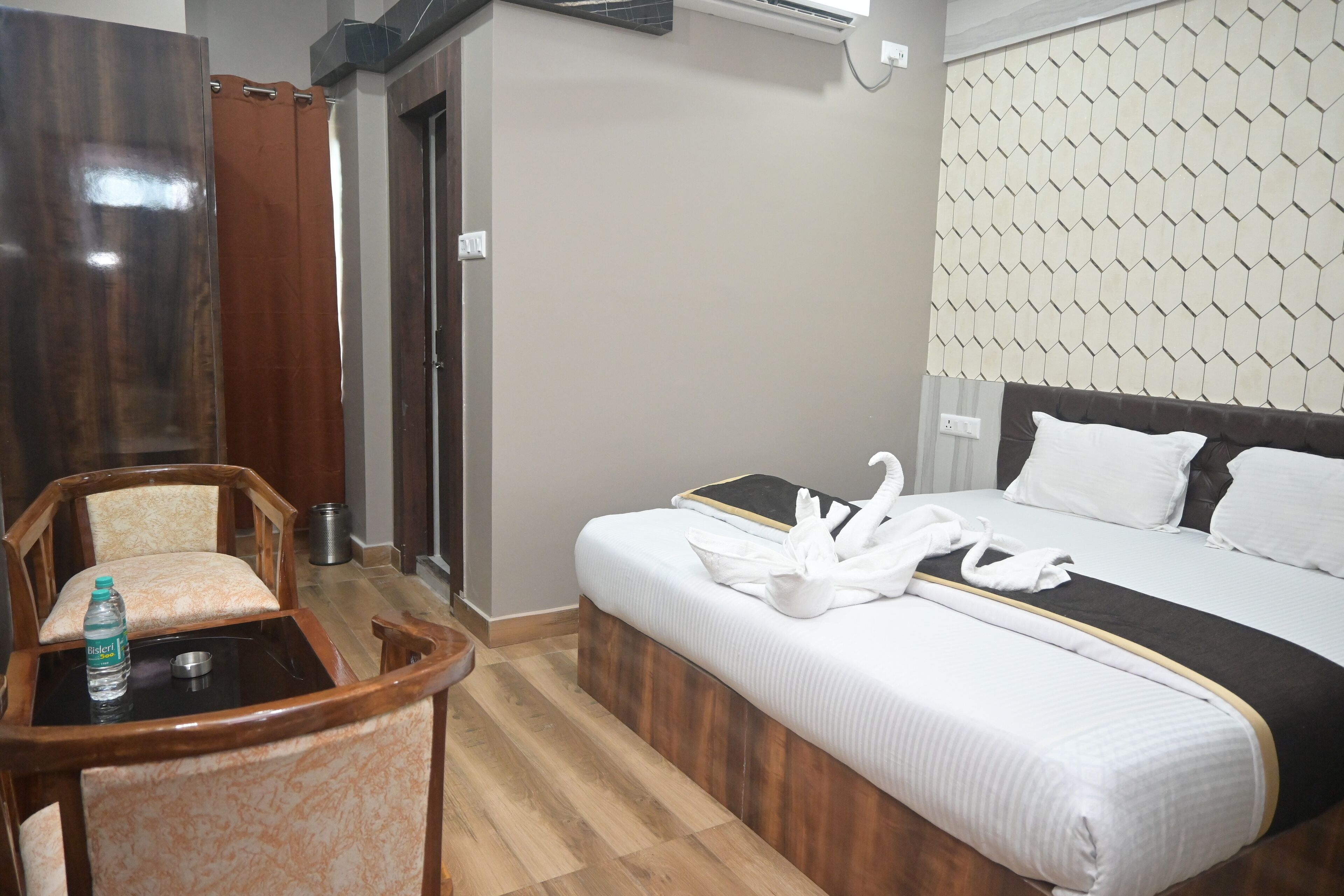 A hotel room with a bed, a wooden chair, a small table with a bottle, and a wall-mounted air conditioner.
