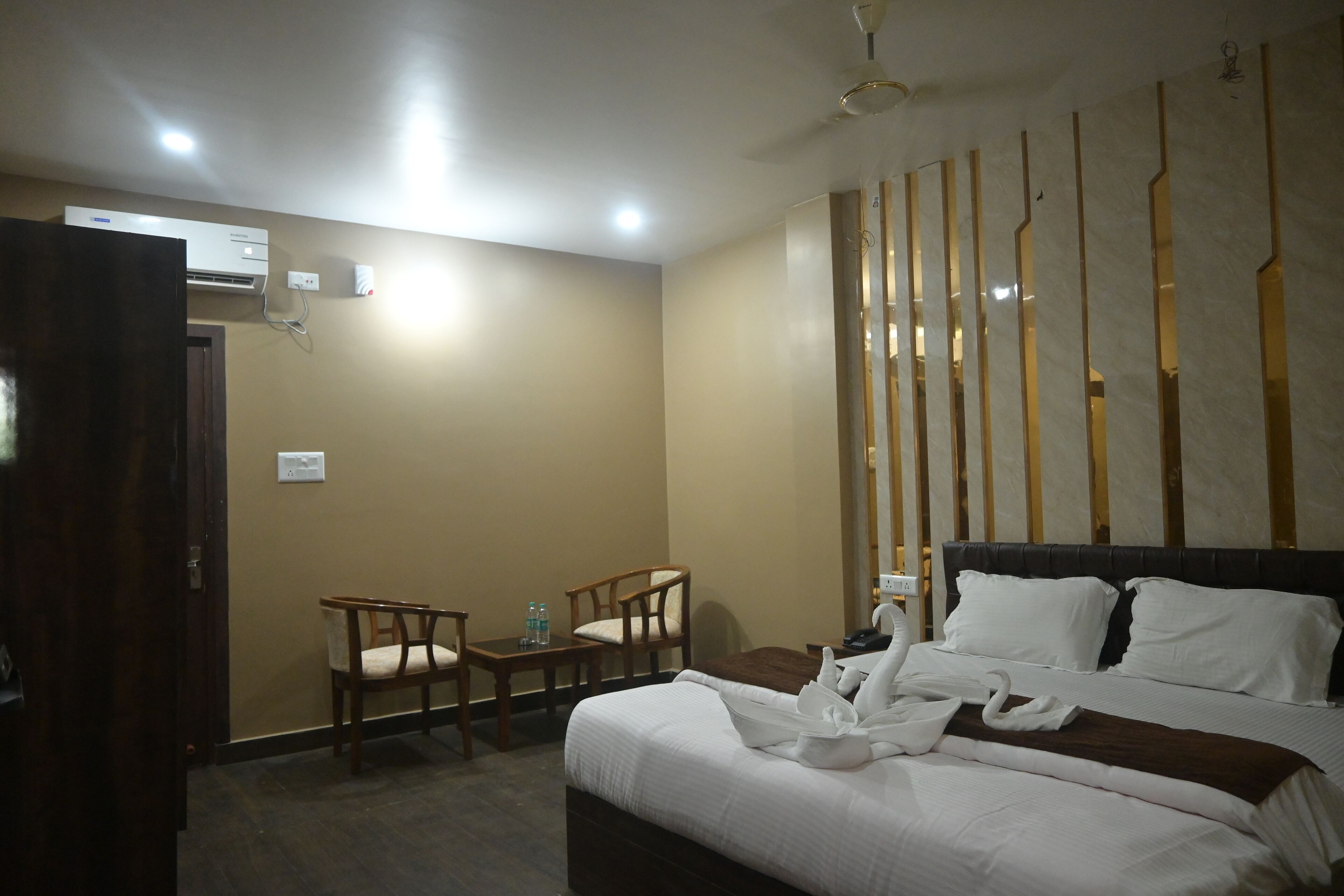A hotel room with a bed, two chairs, a small table with water bottles, an air conditioning unit, and a decorative wall panel.