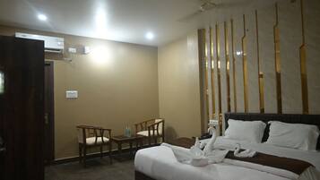 A hotel room with a bed, two chairs, a small table with water bottles, an air conditioning unit, and a decorative wall panel.