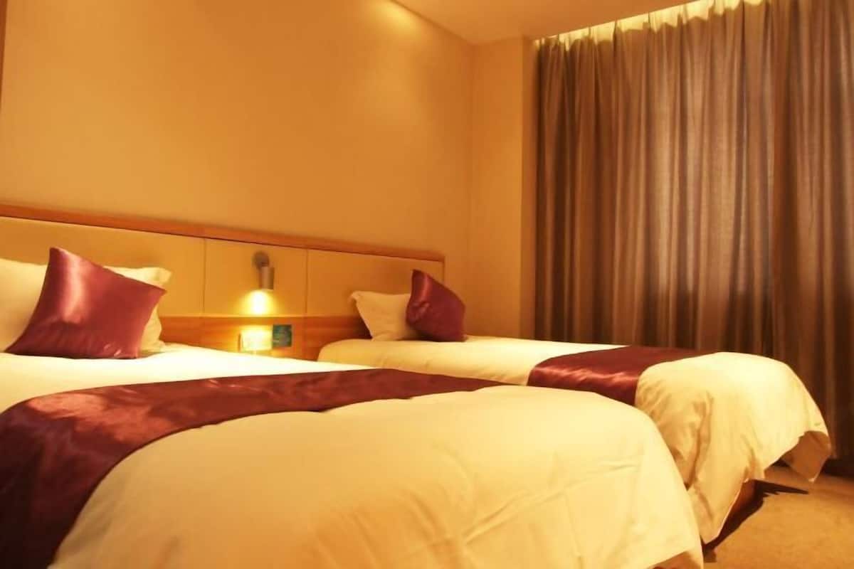 A hotel room with two beds, a wooden headboard, a bedside table with a lamp, and a large window with curtains.