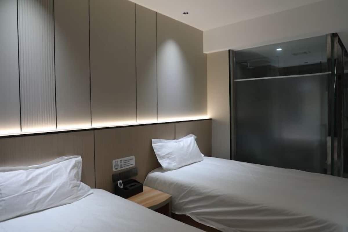A hotel room with two single beds, a bedside table, a small desk, and a glass shower enclosure.
